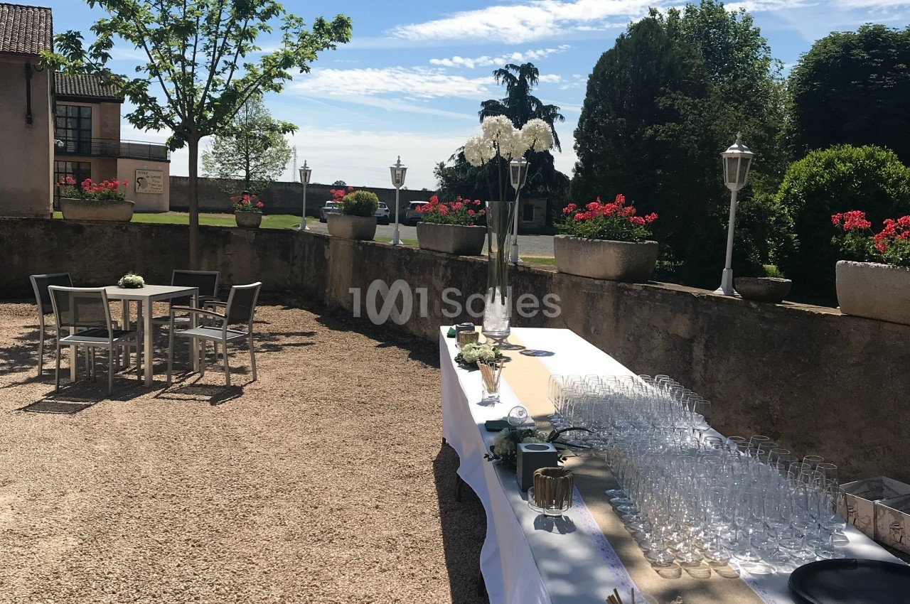 Table dressée avec des verres sur une terrasse ensoleillée, entourée de plantes en pot et de mobilier de jardin.