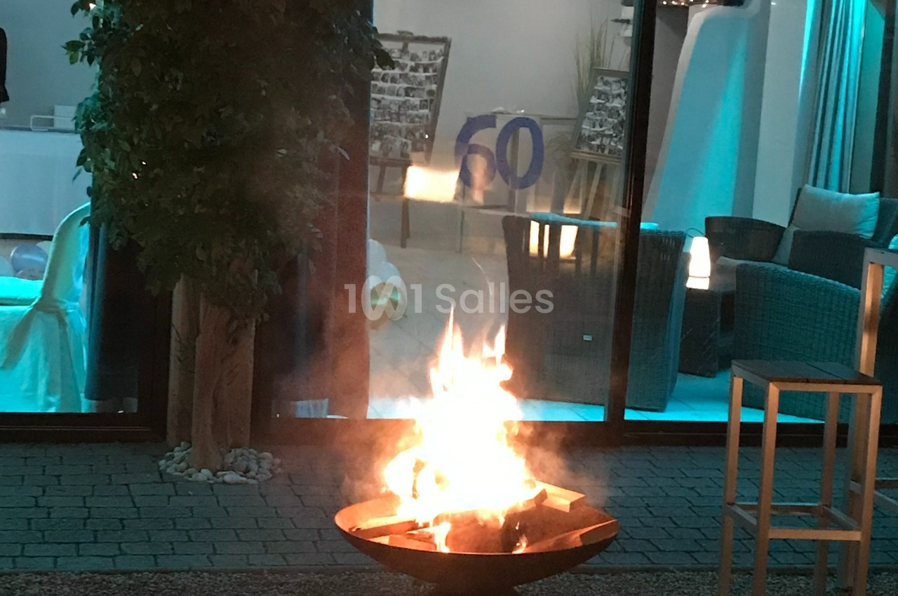 Brasero allumé sur une terrasse en soirée, devant une baie vitrée éclairée et un décor intérieur.