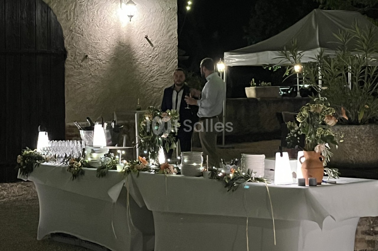 Table de buffet éclairée en extérieur, décorée de fleurs, avec deux hommes discutant à proximité sous un éclairage tamisé.