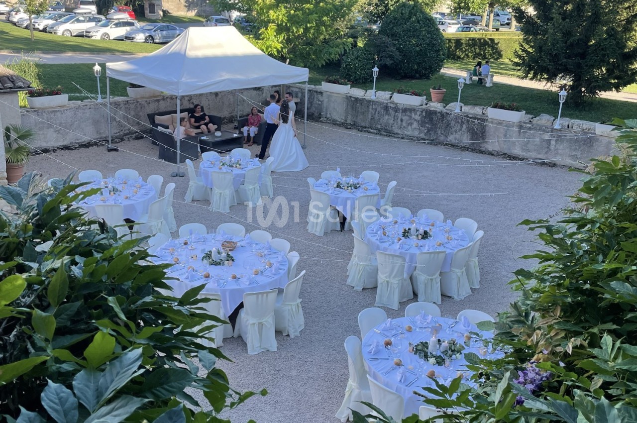 Vue d'une réception en plein air avec tables rondes dressées et un couple en tenue de mariage près d'une tente.