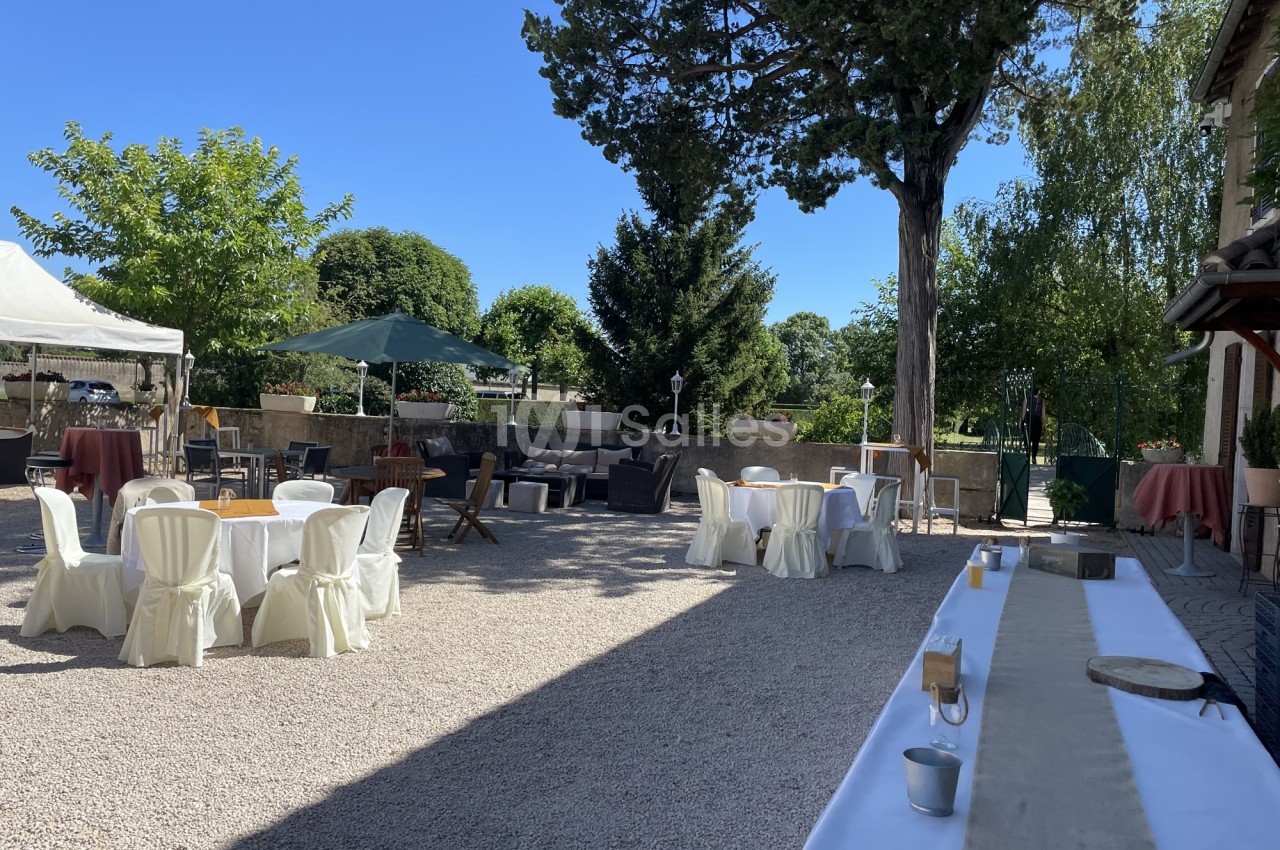 Cour extérieure aménagée pour un événement, avec tables rondes dressées, chaises couvertes et parasols sous un ciel dégagé.