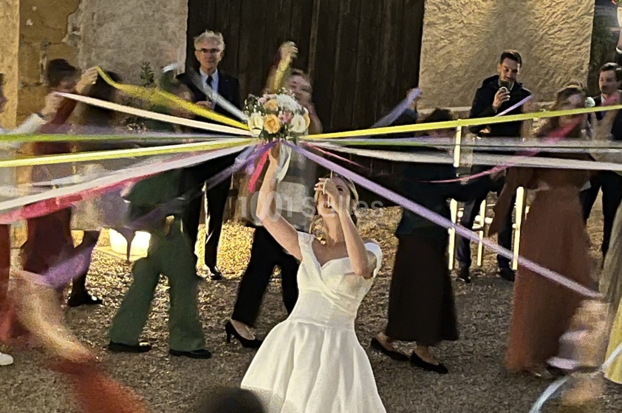 Une mariée en robe blanche tient un bouquet entouré de rubans colorés, avec des invités en arrière-plan.