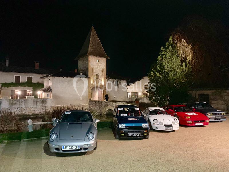 Alignement de voitures anciennes devant un bâtiment historique éclairé de nuit, entouré de végétation.