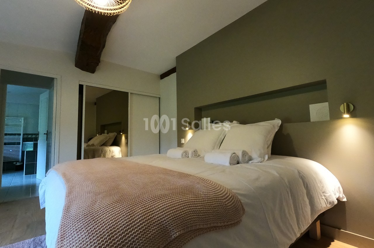 Lit double avec couverture beige, draps blancs et serviettes pliées, dans une chambre lumineuse avec poutre apparente.