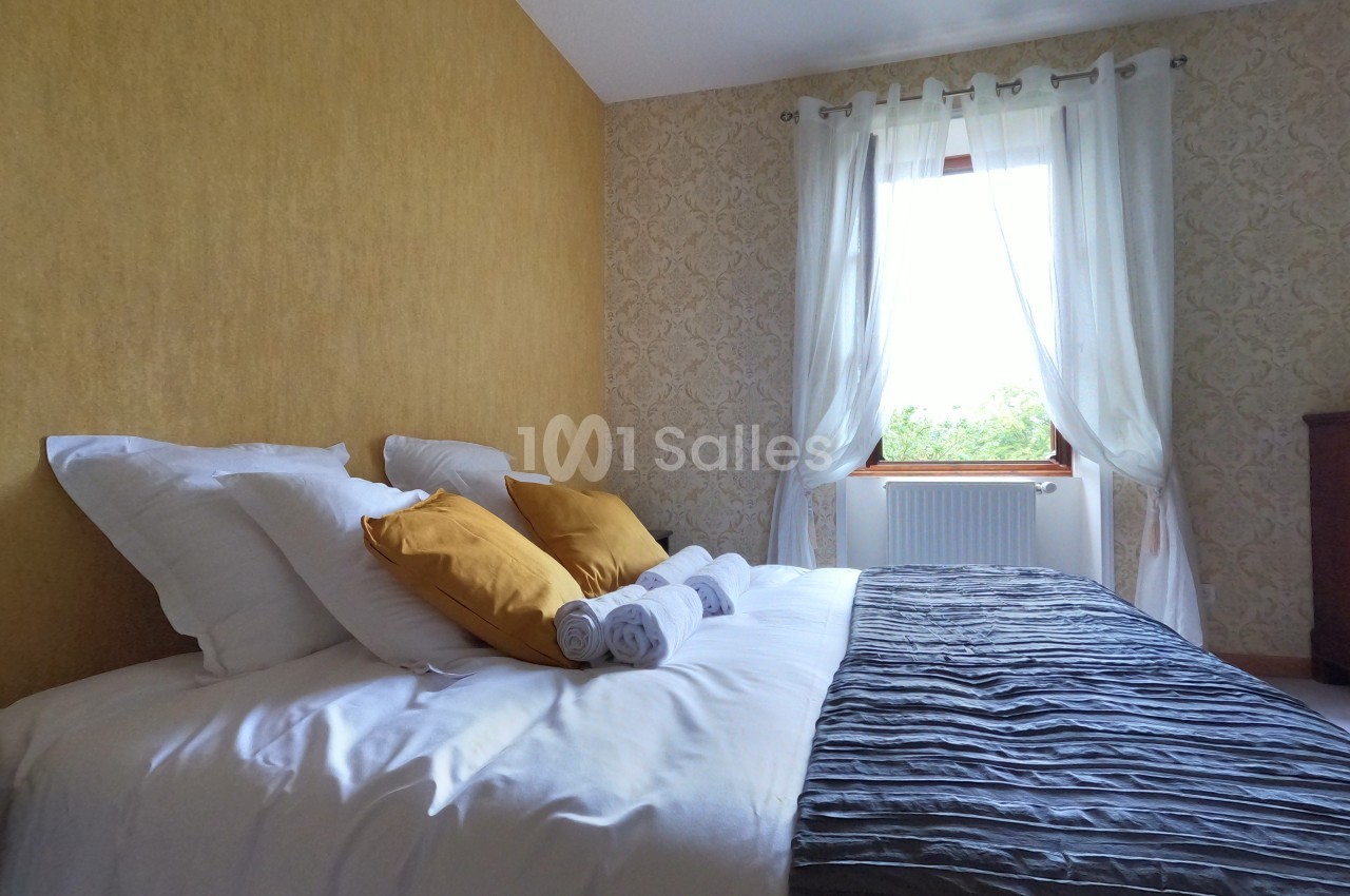 Lit double avec draps blancs, coussins jaunes, serviettes pliées, dans une chambre lumineuse aux murs jaunes et beige.