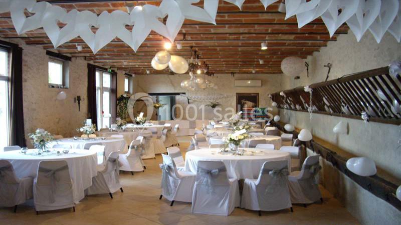 Salle de réception décorée pour un événement, avec tables rondes habillées de nappes blanches et guirlandes suspendues.
