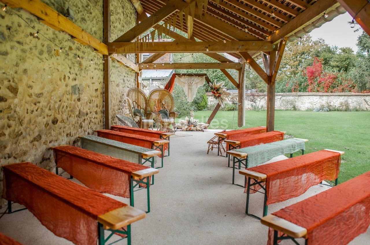 Espace de cérémonie en plein air sous une pergola en bois, avec bancs décorés de tissus colorés et vue sur un jardin.