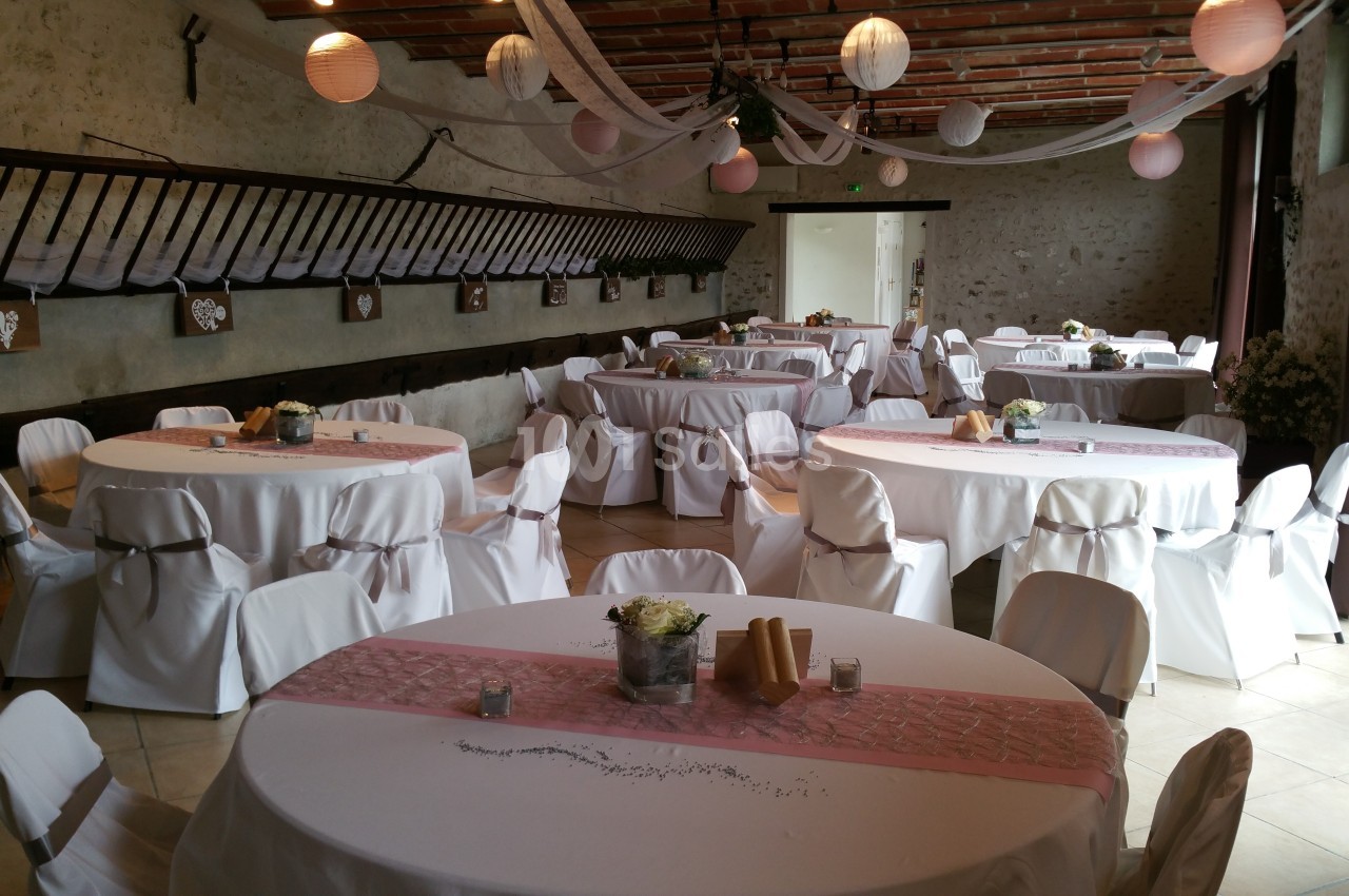 Salle de réception décorée avec des tables rondes, nappes blanches, chemins de table roses et lanternes suspendues.