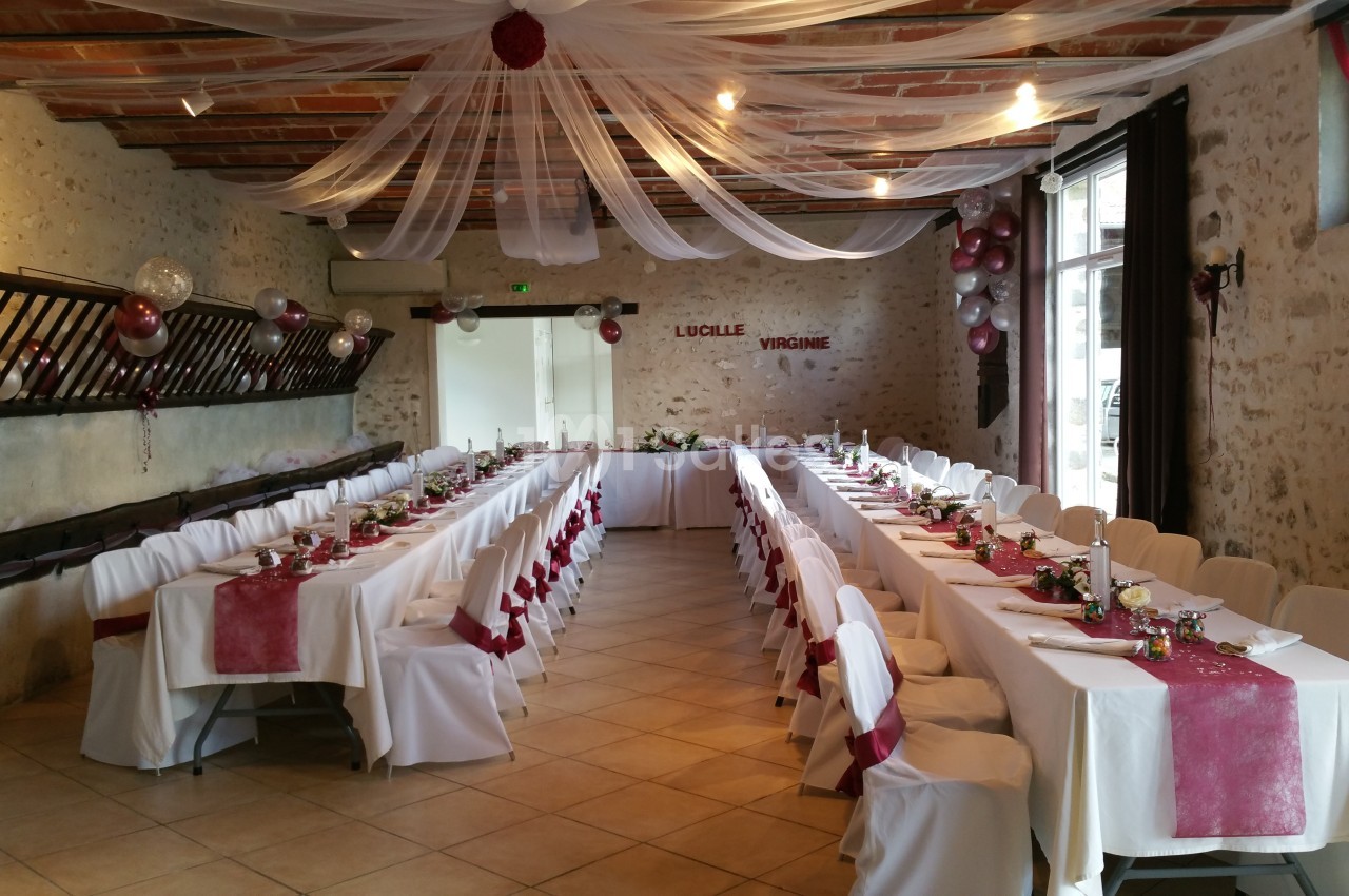Salle de réception décorée avec des tables alignées, nappes blanches, chemins de table rouges et guirlandes suspendues.