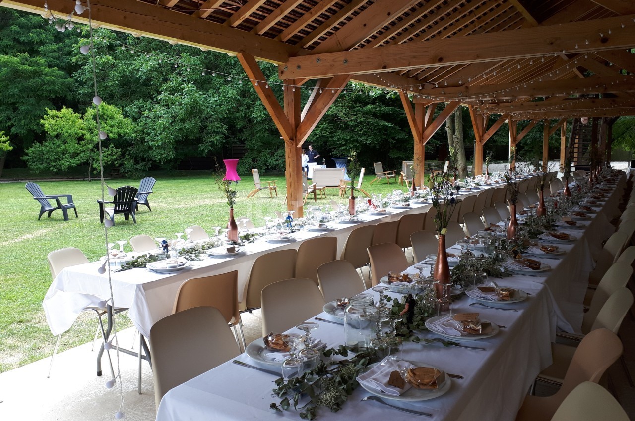 Tables dressées pour un repas en extérieur sous une pergola en bois, avec vue sur un jardin verdoyant.