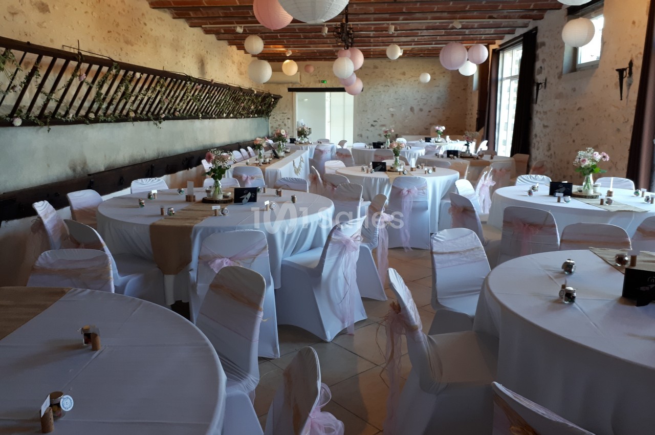 Salle de réception décorée avec des tables rondes, nappes blanches, chaises ornées de rubans roses et lanternes suspendues.