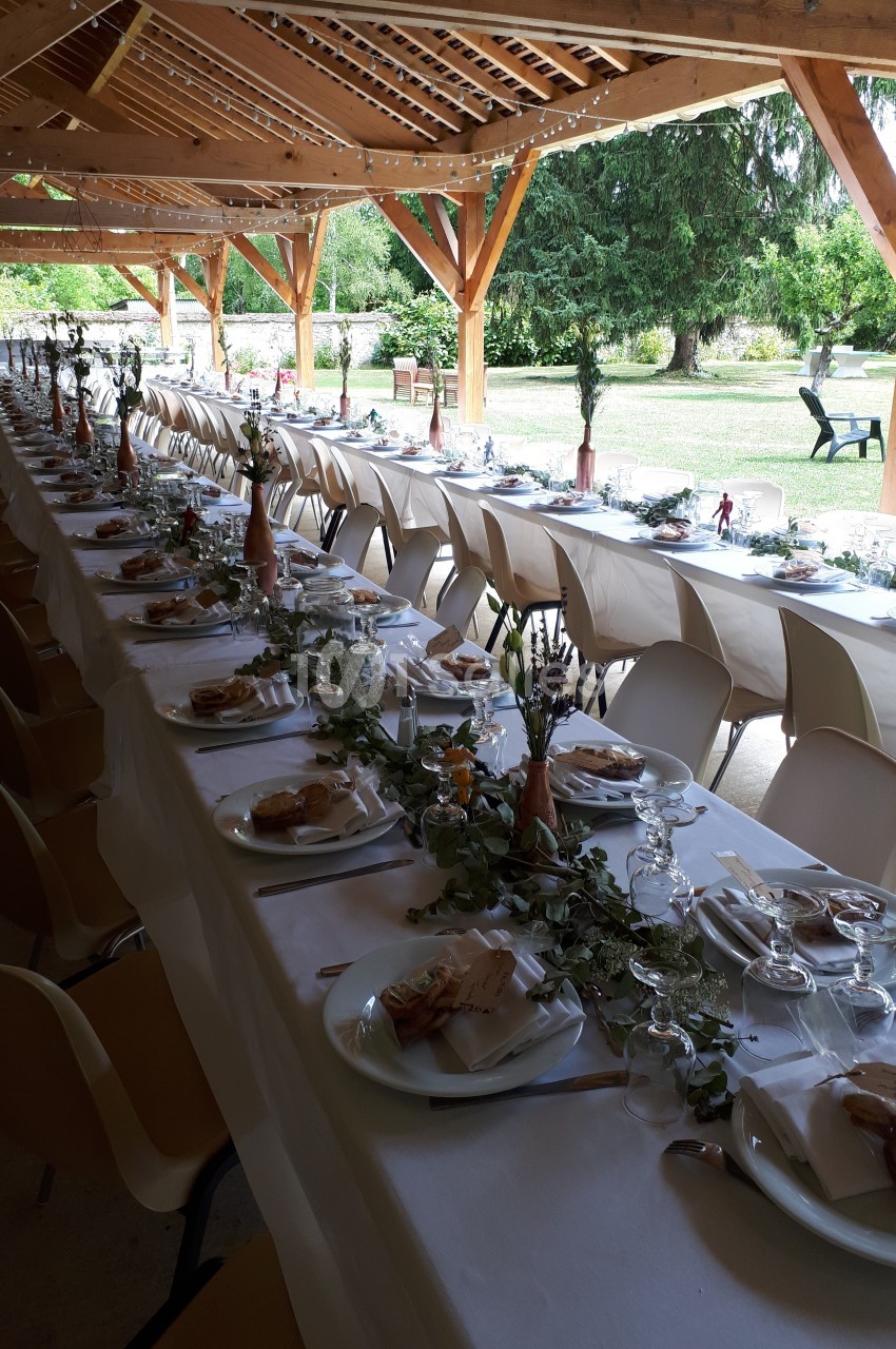 Tables longues décorées pour un repas festif sous une structure en bois, avec vue sur un jardin verdoyant.