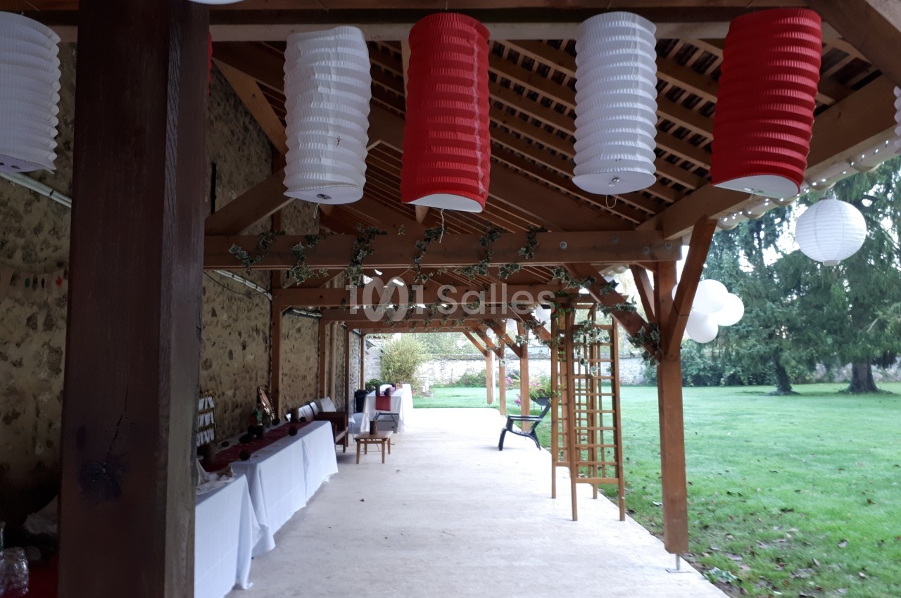 Pergola en bois décorée de lanternes rouges et blanches, tables dressées sur le côté, donnant sur un jardin verdoyant.