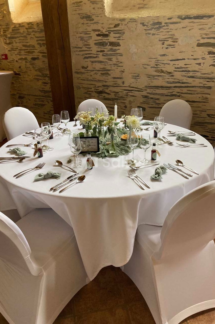 Table ronde dressée pour un repas, avec nappes blanches, couverts, verres, serviettes et centre de table floral.
