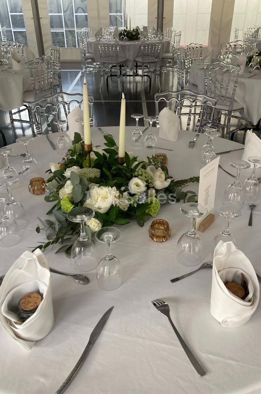 Table ronde dressée avec une nappe blanche, décorée de bougies, fleurs blanches et vertes, entourée de chaises transparentes.