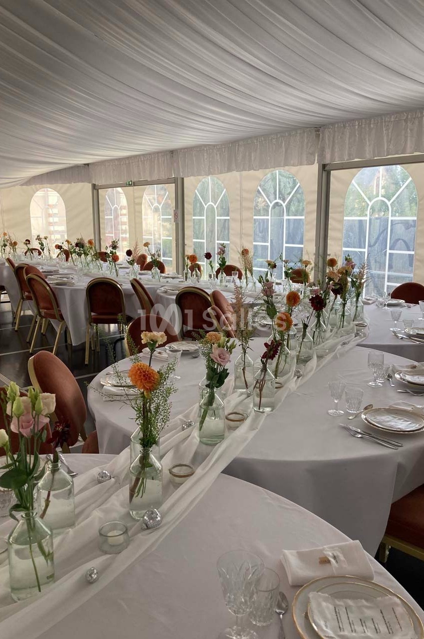 Salle de réception décorée avec des tables rondes, nappes blanches et bouquets de fleurs dans des vases en verre.