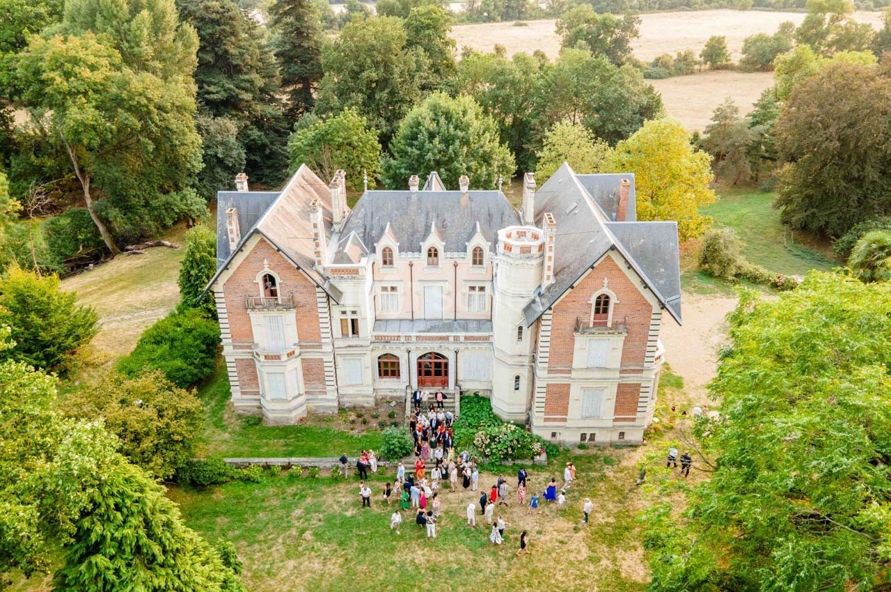 Vue aérienne d'un château ancien entouré de verdure, avec un groupe de personnes rassemblées devant l'entrée.