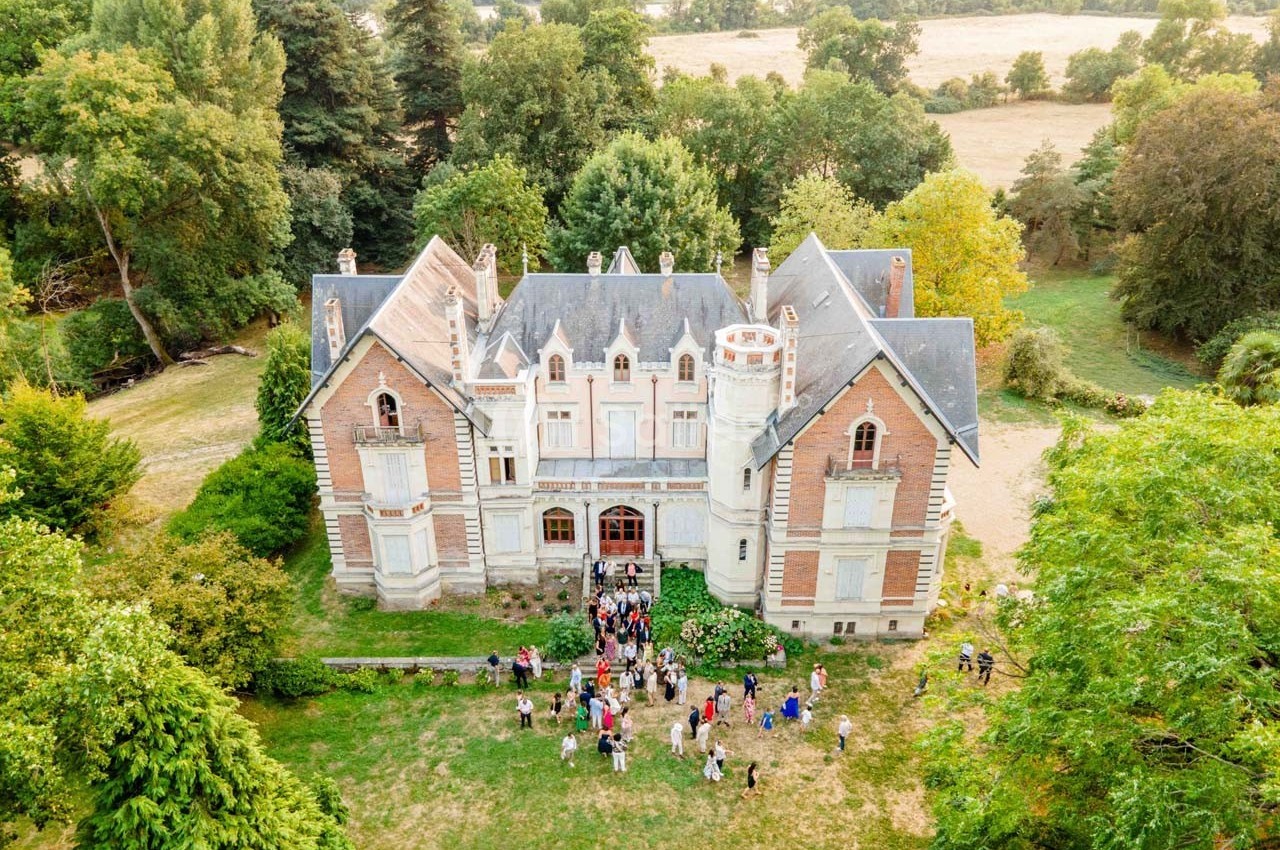 Vue aérienne d'un château entouré de verdure, avec un rassemblement de personnes devant l'entrée principale.