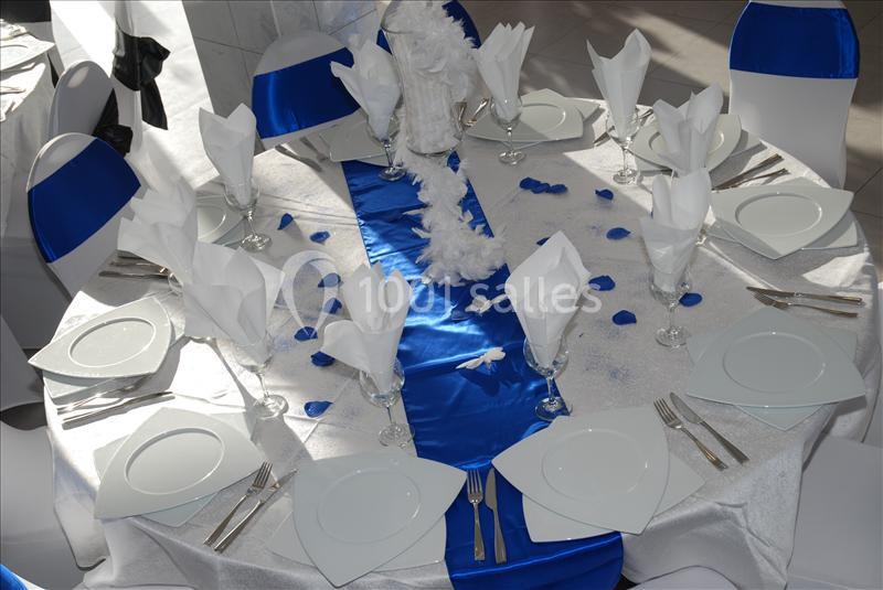 Table dressée pour un événement, nappée de blanc et bleu, avec serviettes pliées, vaisselle blanche et décorations.