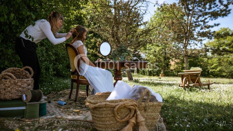 Une femme en robe blanche se fait coiffer à l'extérieur, assise à une table dans un jardin verdoyant.