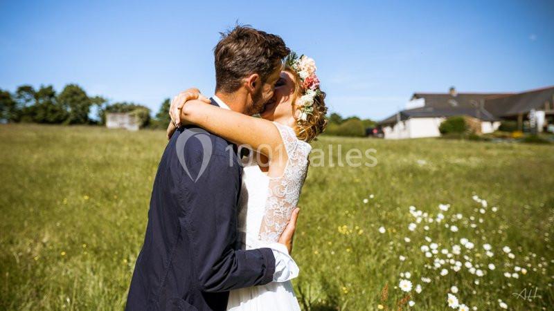 Un couple s'embrasse dans un champ fleuri sous un ciel bleu, près d'une maison en arrière-plan.