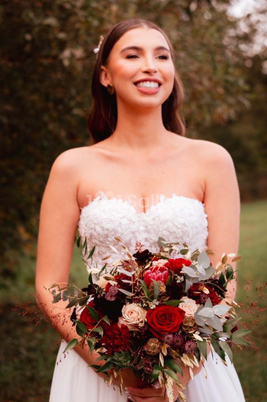 Une femme en robe de mariée tient un bouquet de fleurs rouges et blanches dans un cadre naturel.