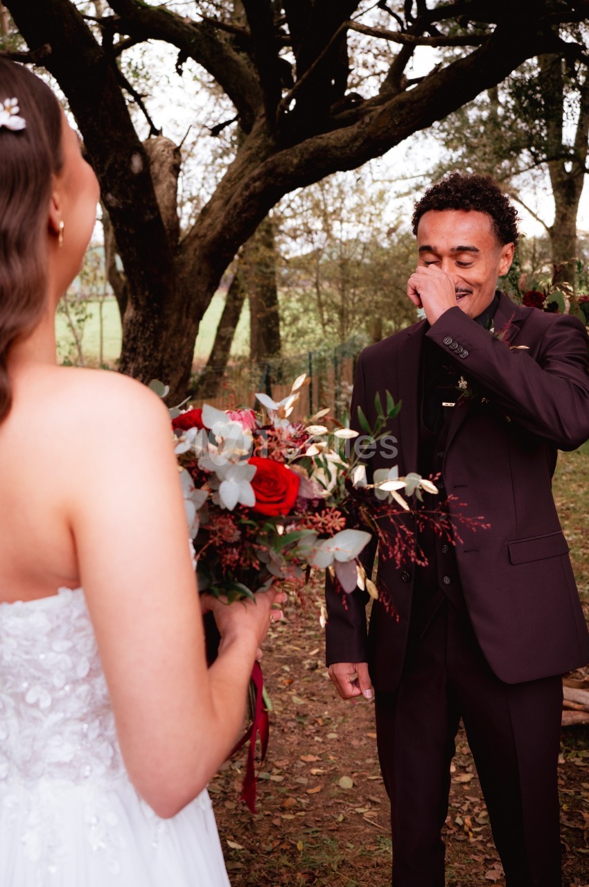 Un homme ému en costume sombre regarde une femme en robe blanche tenant un bouquet de fleurs rouges, dans un cadre boisé.