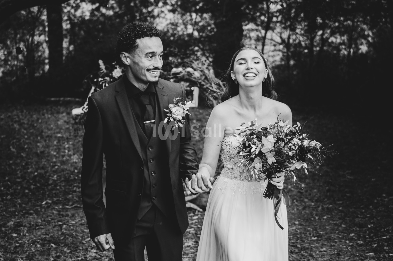 Un couple souriant marche main dans la main dans un cadre extérieur boisé, vêtus de tenues de mariage.