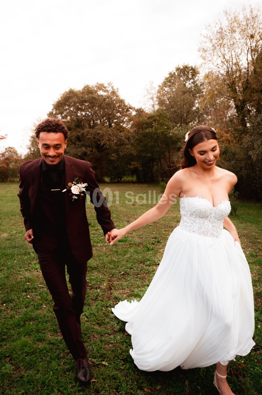 Un couple en tenue de mariage marche main dans la main dans un champ verdoyant sous un ciel légèrement nuageux.