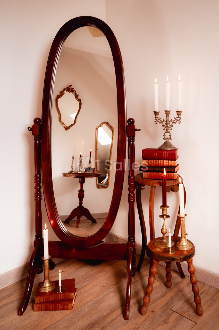 Grand miroir ovale sur pied entouré de bougies, livres anciens et meubles en bois dans un intérieur chaleureux.