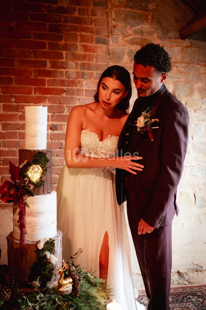 Un couple en tenue de mariage observe un gâteau à étages décoré de fleurs, dans un décor rustique en briques.