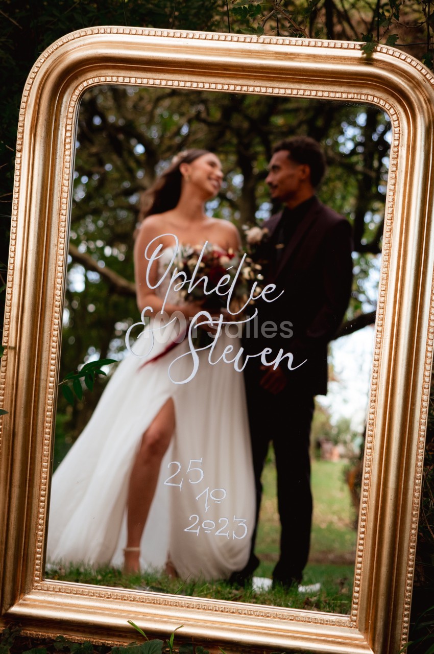 Un couple souriant se reflète dans un miroir doré entouré de végétation, avec des inscriptions manuscrites sur la surface.