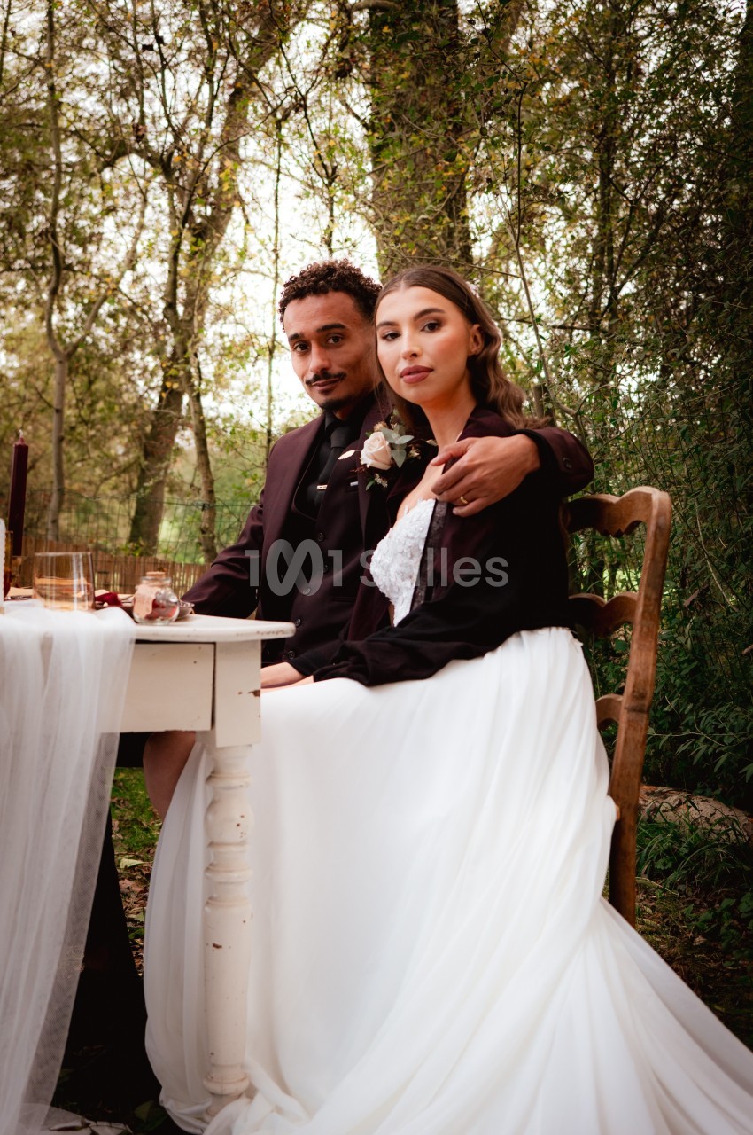 Un couple assis à une table décorée dans un cadre boisé, la femme porte une robe blanche élégante.