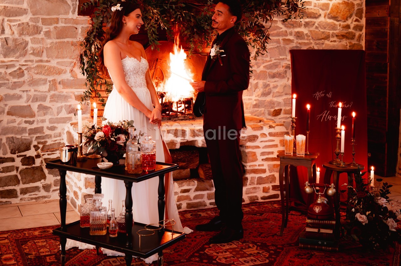 Un couple en tenue de mariage se tient devant une cheminée allumée, entouré de décorations chaleureuses et élégantes.