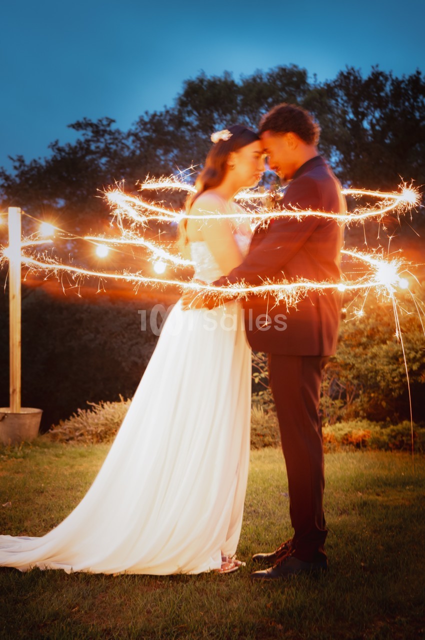 Un couple en tenue de mariage s'enlace dans un jardin, entouré d'étincelles lumineuses au crépuscule.