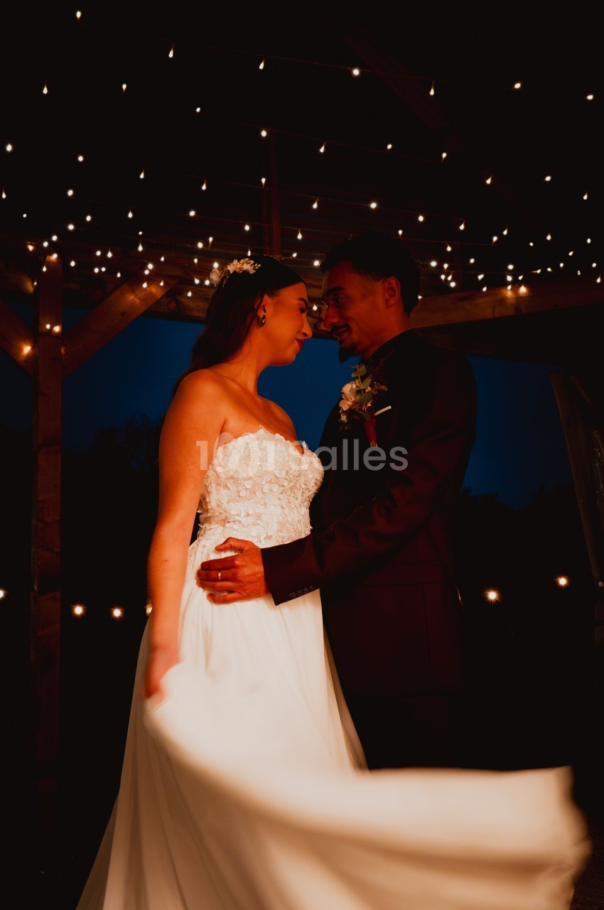 Un couple en tenue de mariage se regarde sous une pergola illuminée de guirlandes lumineuses.