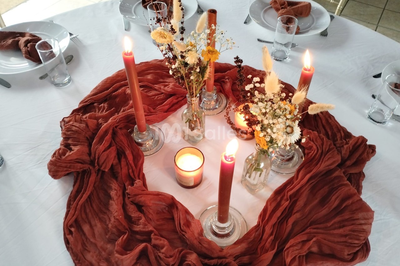 Centre de table avec bougies allumées, fleurs séchées et tissu rouge sur une nappe blanche.