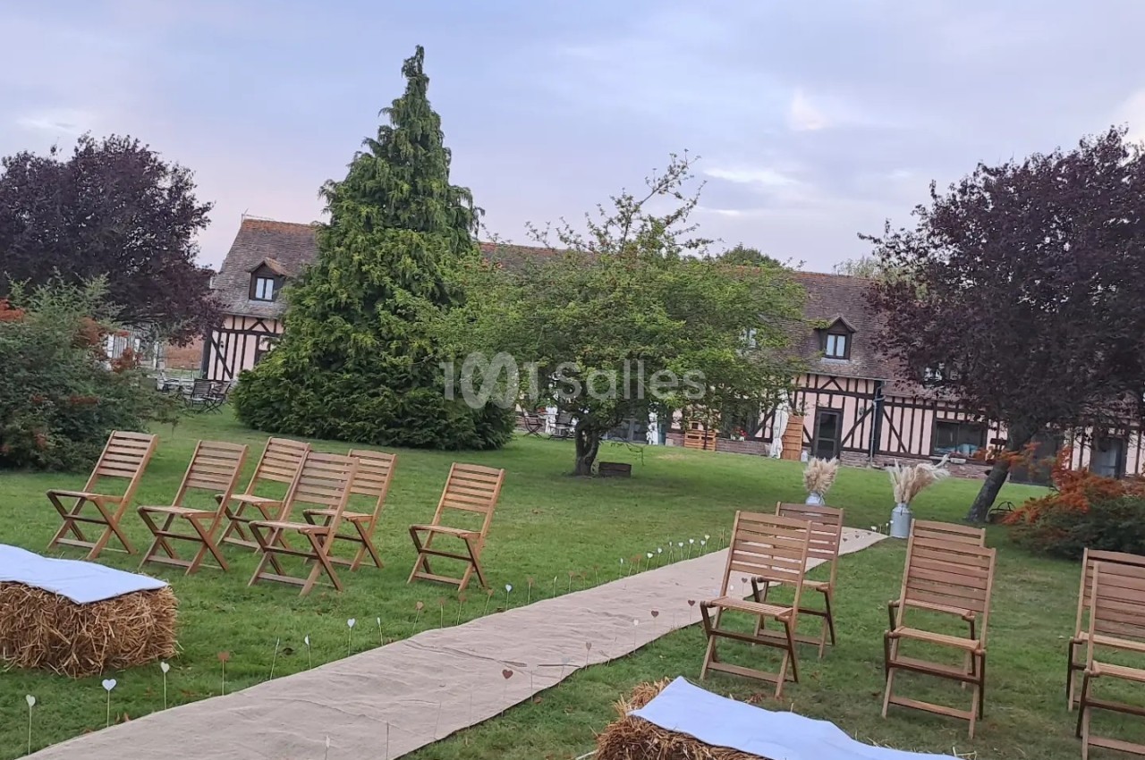 Allée centrale en tissu bordée de chaises en bois et bottes de paille, dans un jardin verdoyant devant une maison à…