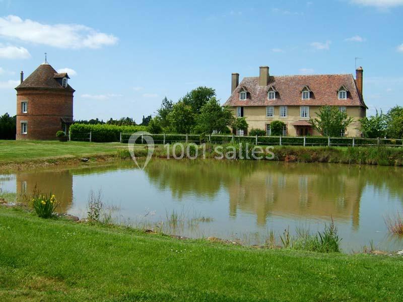 Maison de campagne avec un étang au premier plan, entourée de verdure et d'un petit bâtiment en briques.