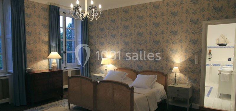 Chambre avec lit double, murs à motif floral, fenêtres avec rideaux bleus, et salle de bain attenante éclairée.