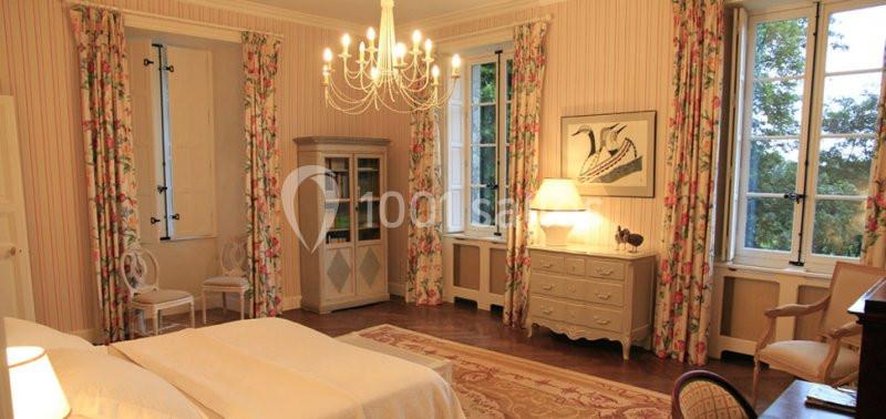 Chambre lumineuse avec un lit double, des rideaux fleuris, un lustre, des meubles classiques et des fenêtres donnant sur la…